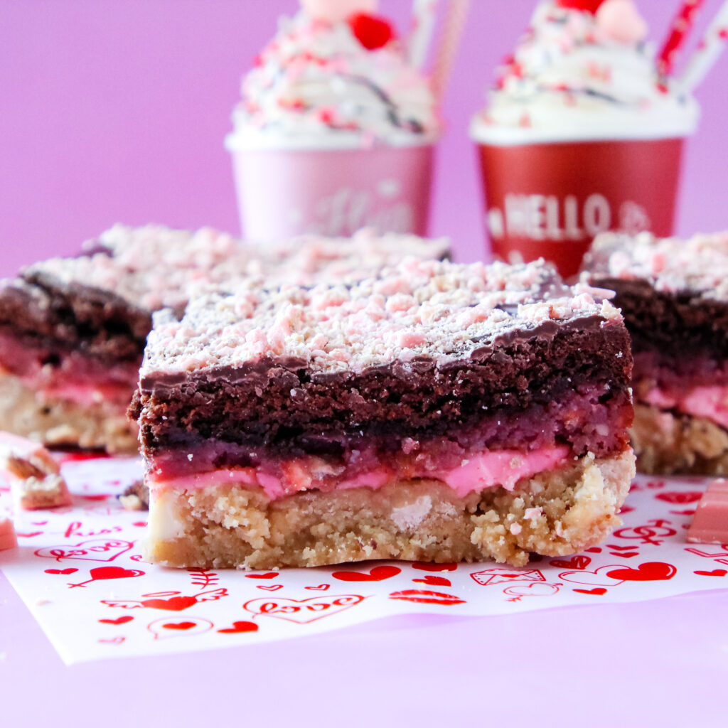 A stack of Raspberry Bombshell Sweet Stacks with layers of blondie, gooey raspberry chocolate cake, and pink cream cheese frosting, drizzled with milk chocolate and topped with crushed raspberry candy.