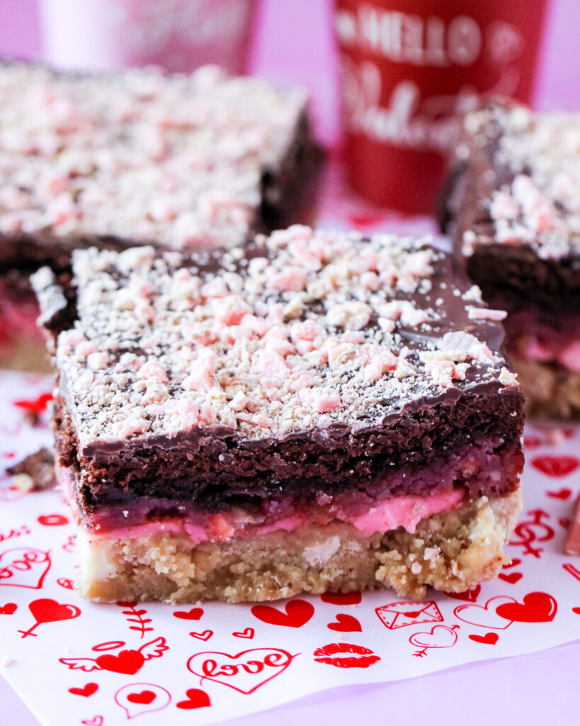 A stack of Raspberry Bombshell Sweet Stacks with layers of blondie, gooey raspberry chocolate cake, and pink cream cheese frosting, drizzled with milk chocolate and topped with crushed raspberry candy.