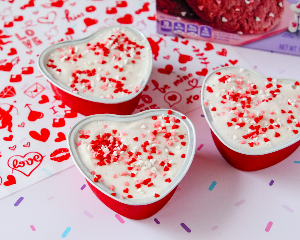 Heart-shaped red velvet cookie cups filled with ice cream, topped with colorful sprinkles, served in mini containers—perfect for a festive family treat.