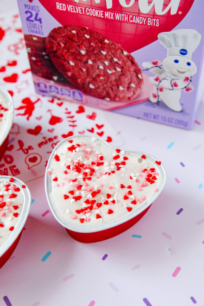 Heart-shaped red velvet cookie cups filled with ice cream, topped with colorful sprinkles, served in mini containers—perfect for a festive family treat.