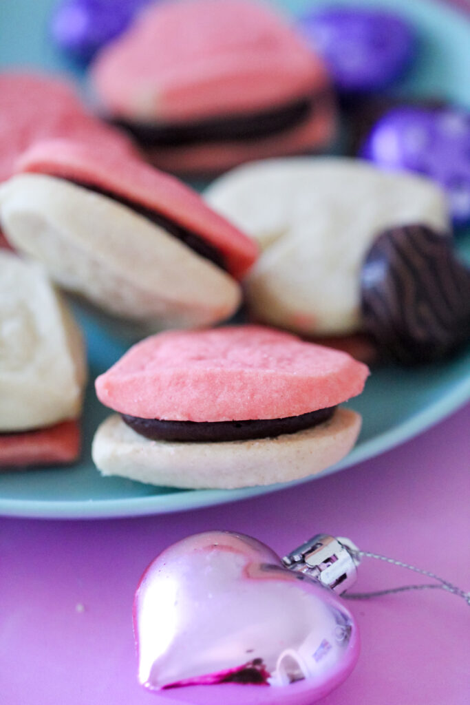 A plate of heart-shaped cookie sandwiches filled with creamy fudge frosting, perfect for Valentine’s Day, a girls' night in, or a rom-com movie marathon. The cookies are pink and golden, with rich chocolate frosting peeking between the layers.