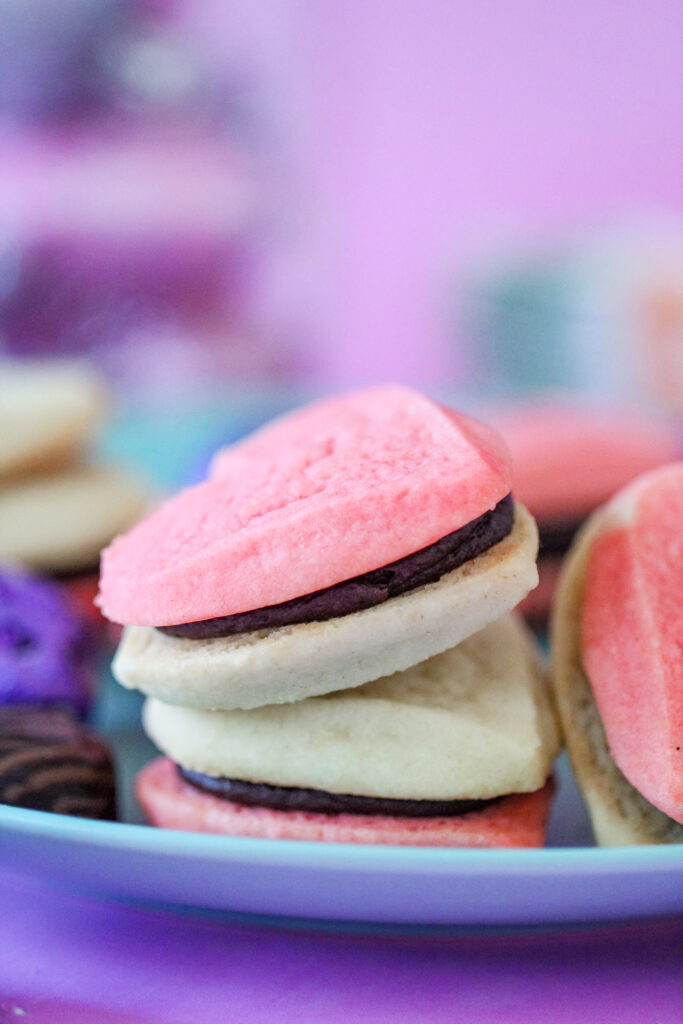 A plate of heart-shaped cookie sandwiches filled with creamy fudge frosting, perfect for Valentine’s Day, a girls' night in, or a rom-com movie marathon. The cookies are pink and golden, with rich chocolate frosting peeking between the layers.