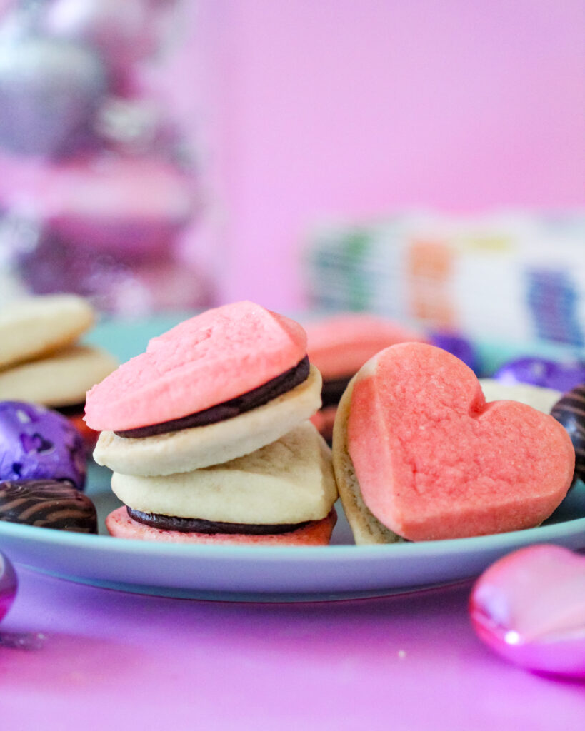 A plate of heart-shaped cookie sandwiches filled with creamy fudge frosting, perfect for Valentine’s Day, a girls' night in, or a rom-com movie marathon. The cookies are pink and golden, with rich chocolate frosting peeking between the layers.