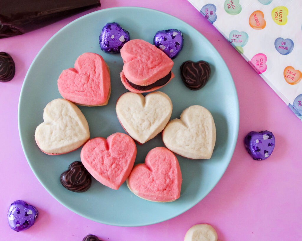 A plate of heart-shaped cookie sandwiches filled with creamy fudge frosting, perfect for Valentine’s Day, a girls' night in, or a rom-com movie marathon. The cookies are pink and golden, with rich chocolate frosting peeking between the layers.