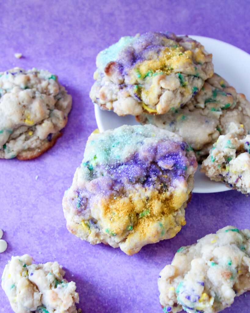 A thick, golden-brown Levain-style cookie topped with festive purple, green, and gold sanding sugar. These cookies have a soft, gooey center with bursts of white chocolate and cream cheese, inspired by the flavors of a traditional Mardi Gras King Cake.