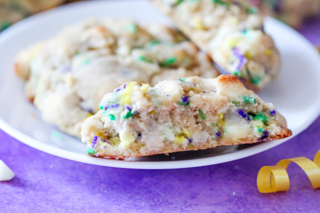 A thick, golden-brown Levain-style cookie topped with festive purple, green, and gold sanding sugar. These cookies have a soft, gooey center with bursts of white chocolate and cream cheese, inspired by the flavors of a traditional Mardi Gras King Cake.