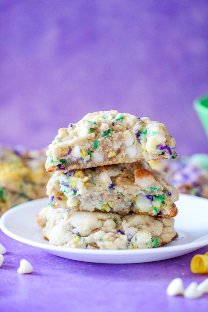 A stack of thick, golden-brown Levain-style cookies topped with festive purple, green, and gold sanding sugar. These cookies have a soft, gooey center with bursts of white chocolate and cream cheese, inspired by the flavors of a traditional Mardi Gras King Cake.