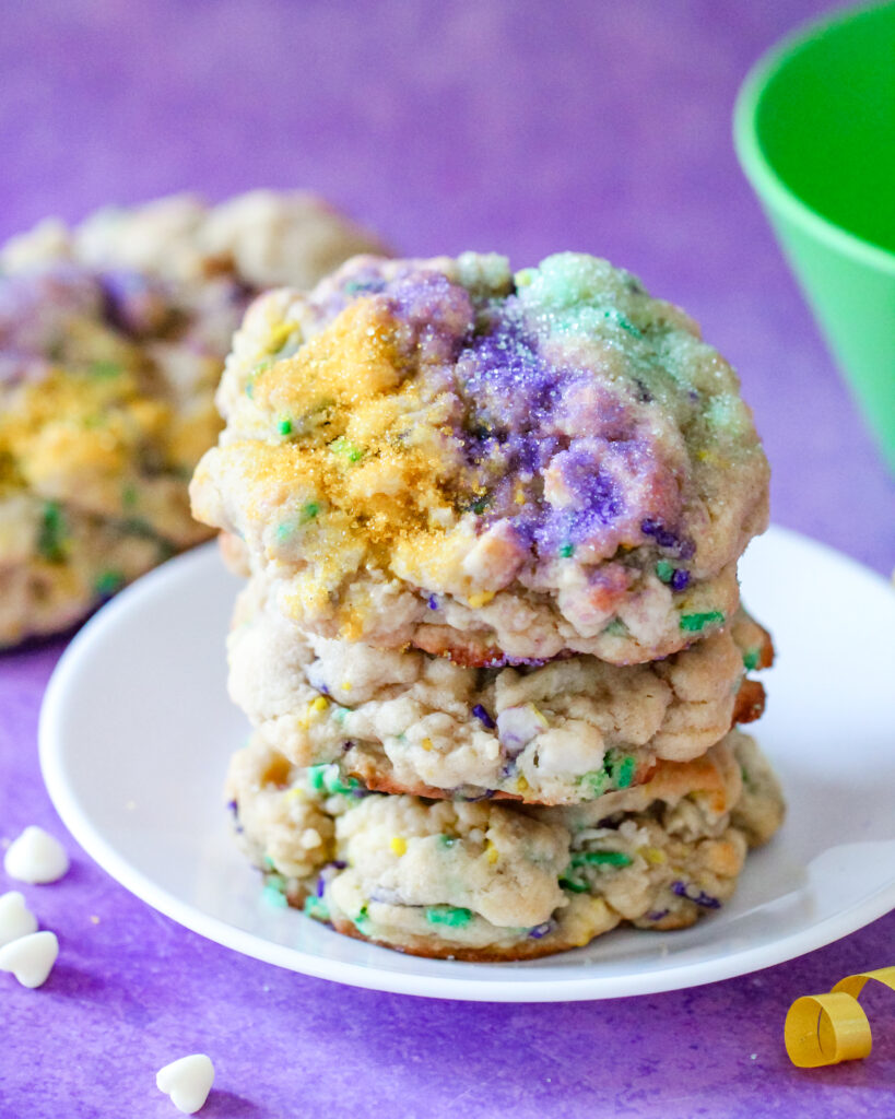 A stack of thick, golden-brown Levain-style cookies topped with festive purple, green, and gold sanding sugar. These cookies have a soft, gooey center with bursts of white chocolate and cream cheese, inspired by the flavors of a traditional Mardi Gras King Cake.