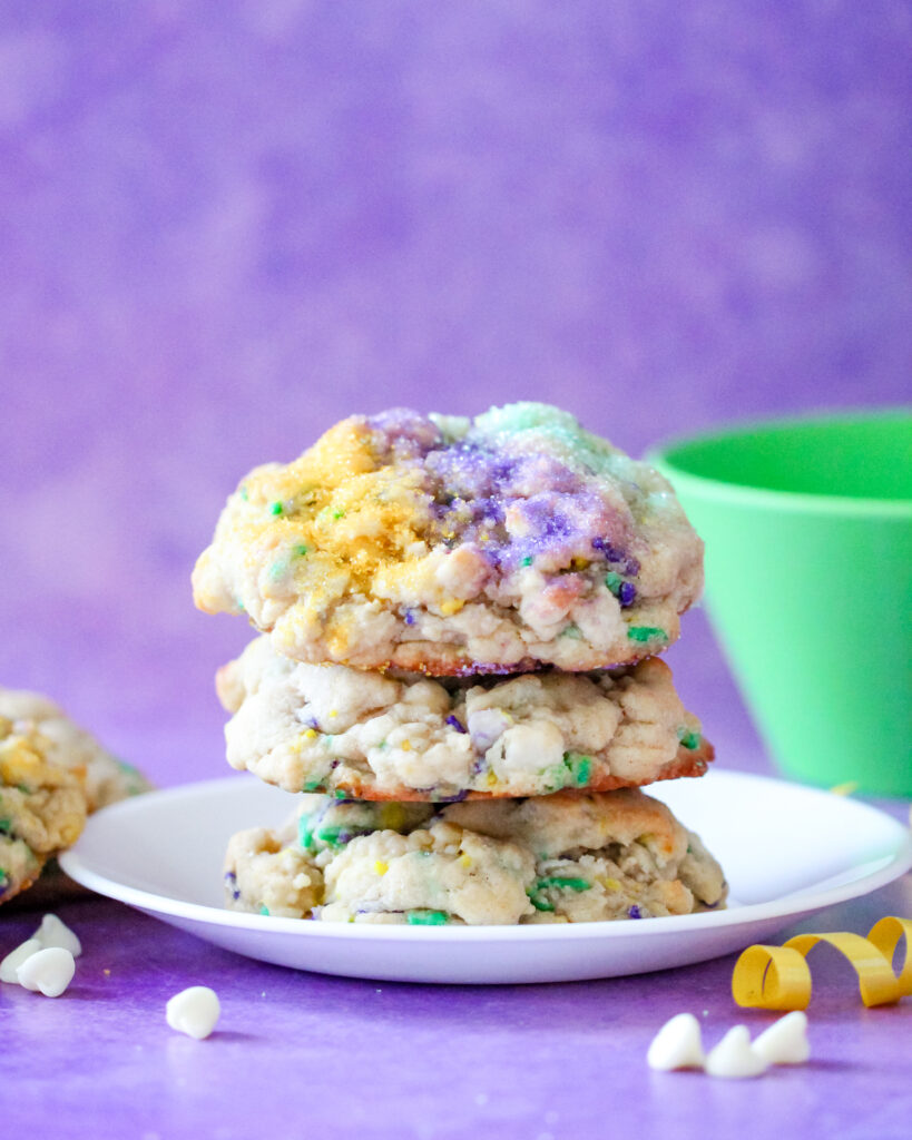 A stack of thick, golden-brown Levain-style cookies topped with festive purple, green, and gold sanding sugar. These cookies have a soft, gooey center with bursts of white chocolate and cream cheese, inspired by the flavors of a traditional Mardi Gras King Cake.