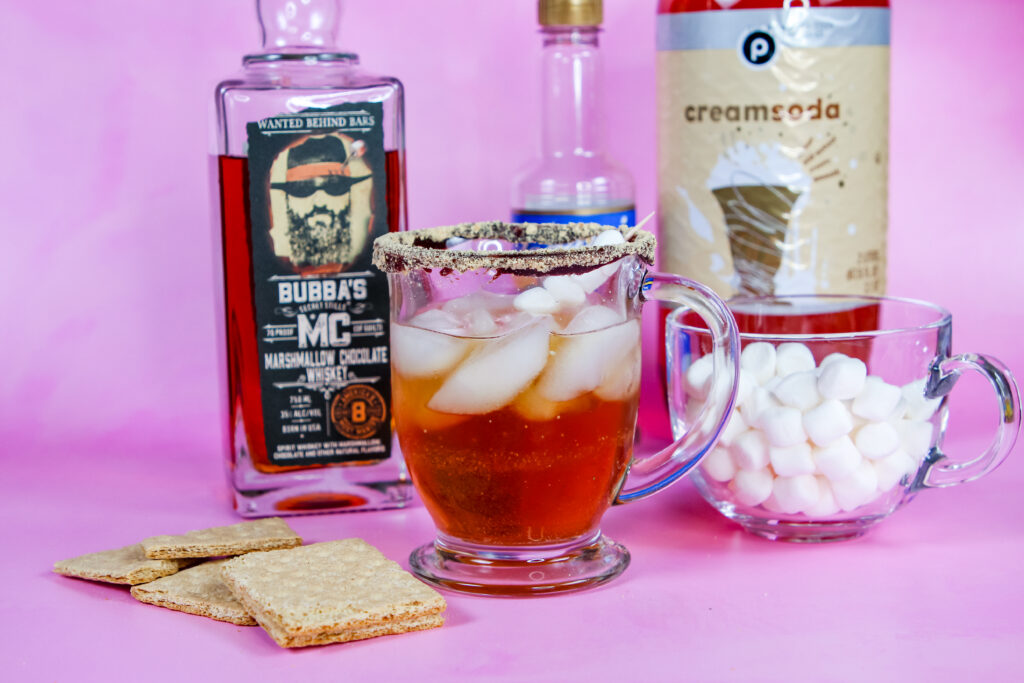 A refreshing S’mores Whiskey Spritzer in a glass with a graham cracker rim, topped with a marshmallow skewer, and filled with a bubbly mix of Bubba’s MC Marshmallow Chocolate Whiskey, vanilla syrup, and cream soda.