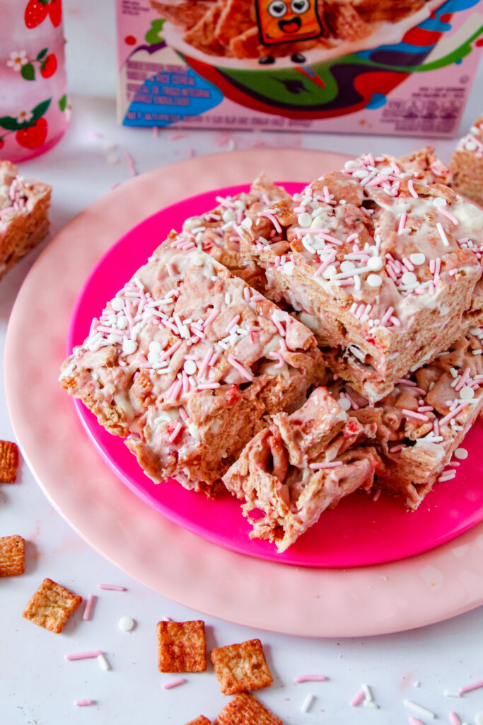 A batch of Strawberry Toast Crunch  Bars, drizzled with pink chocolate and topped with freeze-dried strawberry pieces, cut into squares and stacked on a white plate.