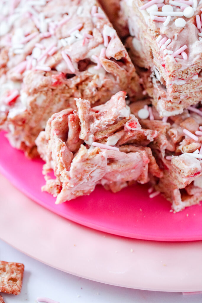 A batch of Strawberry Toast Crunch  Bars, drizzled with pink chocolate and topped with freeze-dried strawberry pieces, cut into squares and stacked on a white plate.