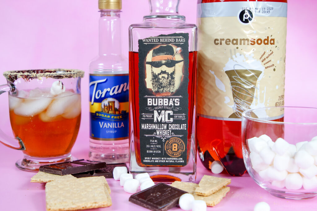 A refreshing S’mores Whiskey Spritzer in a glass with a graham cracker rim, topped with a marshmallow skewer, and filled with a bubbly mix of Bubba’s MC Marshmallow Chocolate Whiskey, vanilla syrup, and cream soda.