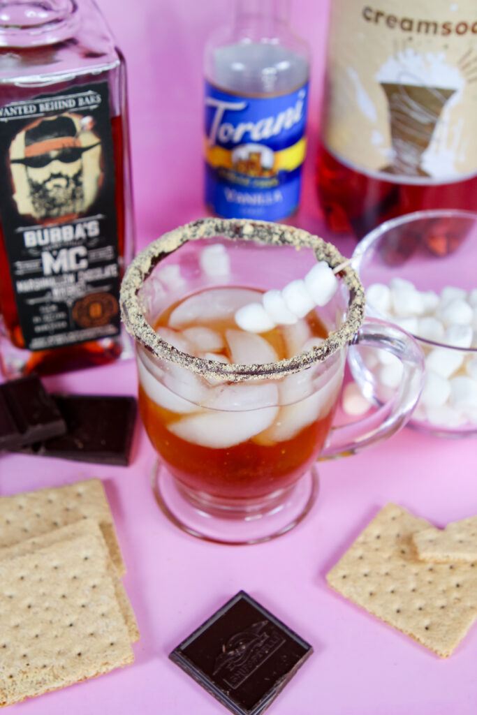 A refreshing S’mores Whiskey Spritzer in a glass with a graham cracker rim, topped with a marshmallow skewer, and filled with a bubbly mix of Bubba’s MC Marshmallow Chocolate Whiskey, vanilla syrup, and cream soda.