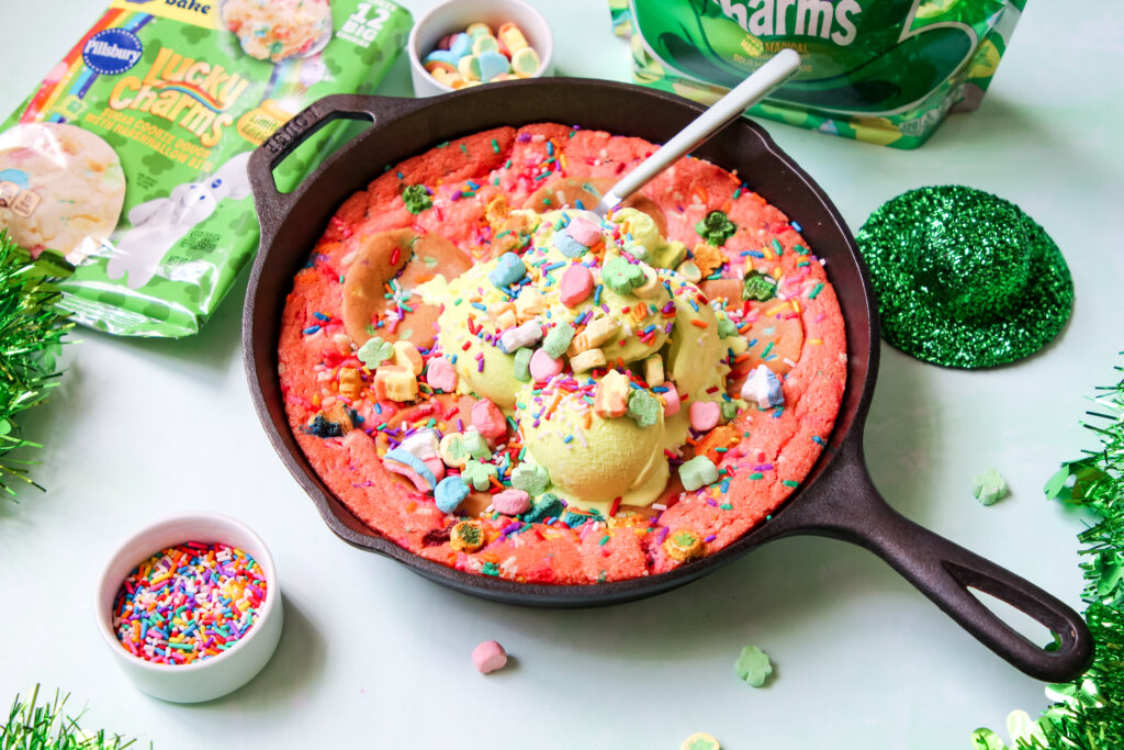 A warm, gooey Lucky Charms Cookie Skillet in a black cast iron pan, topped with green vanilla ice cream, rainbow sprinkles, and Lucky Charms marshmallows.