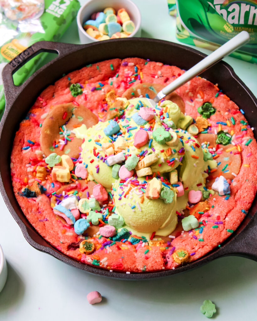 A warm, gooey Lucky Charms Cookie Skillet in a black cast iron pan, topped with green vanilla ice cream, rainbow sprinkles, and Lucky Charms marshmallows.