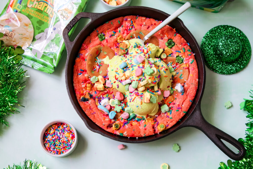 A warm, gooey Lucky Charms Cookie Skillet in a black cast iron pan, topped with green vanilla ice cream, rainbow sprinkles, and Lucky Charms marshmallows.