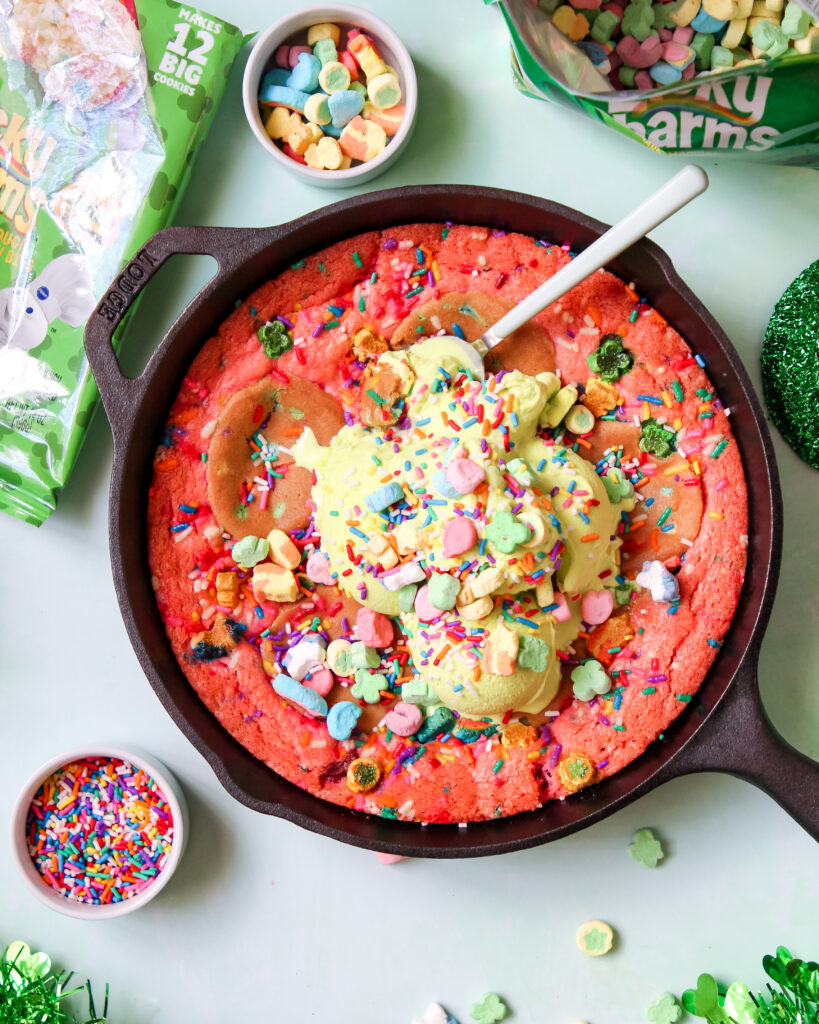 A warm, gooey Lucky Charms Cookie Skillet in a black cast iron pan, topped with green vanilla ice cream, rainbow sprinkles, and Lucky Charms marshmallows.