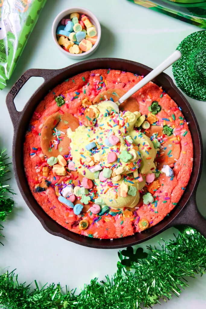 A warm, gooey Lucky Charms Cookie Skillet in a black cast iron pan, topped with green vanilla ice cream, rainbow sprinkles, and Lucky Charms marshmallows.