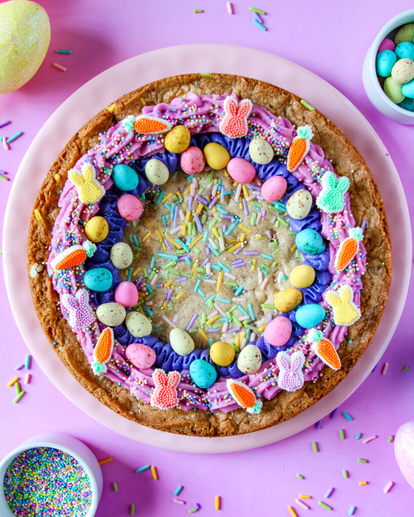 Egg-stravaganza Cookie Cake – a golden chocolate chip cookie cake packed with Easter candies, topped with pastel buttercream swirls and festive sprinkles.