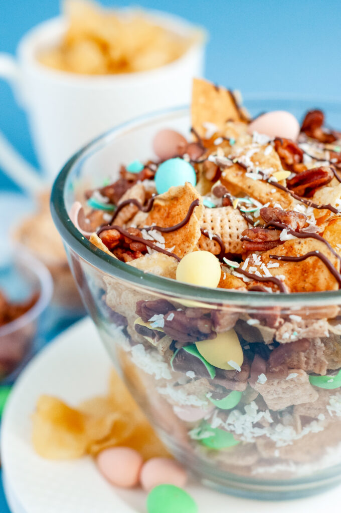 Bowl of garden party snack mix with candied pecans, kettle chips, pastel candies, and a dark chocolate drizzle, sprinkled with flaky sea salt.