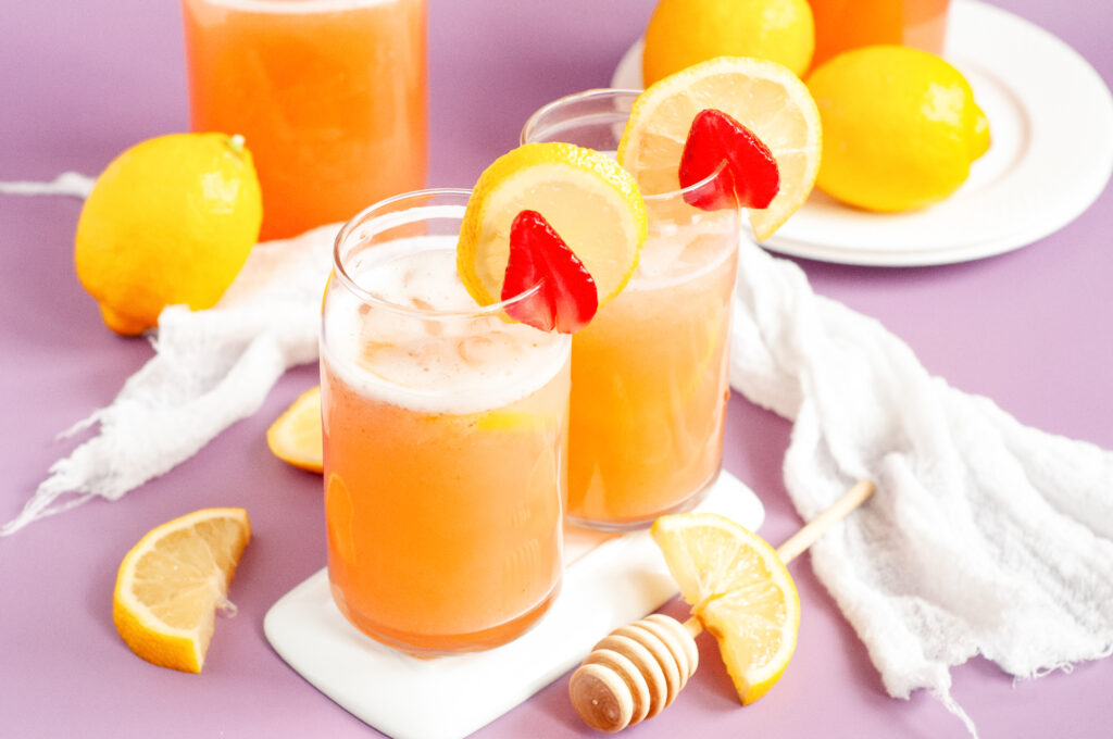 A glass pitcher of fresh strawberry lemonade with floating lemon slices and mint leaves, served over ice with fresh strawberries on the side.