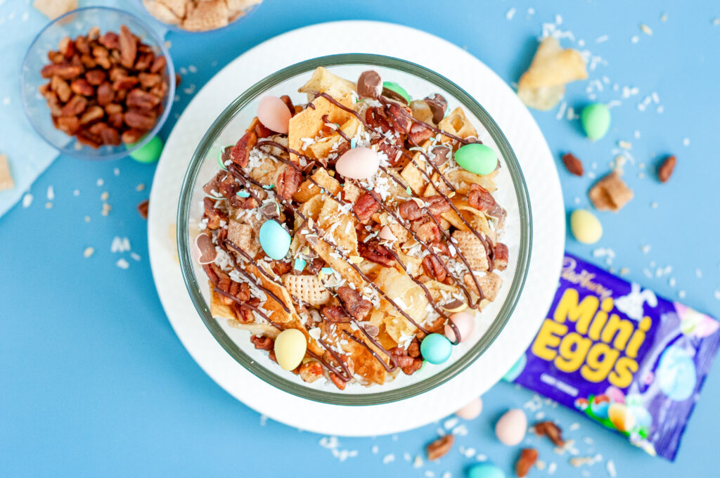 Bowl of garden party snack mix with candied pecans, kettle chips, pastel candies, and a dark chocolate drizzle, sprinkled with flaky sea salt.