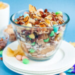 Bowl of garden party snack mix with candied pecans, kettle chips, pastel candies, and a dark chocolate drizzle, sprinkled with flaky sea salt.