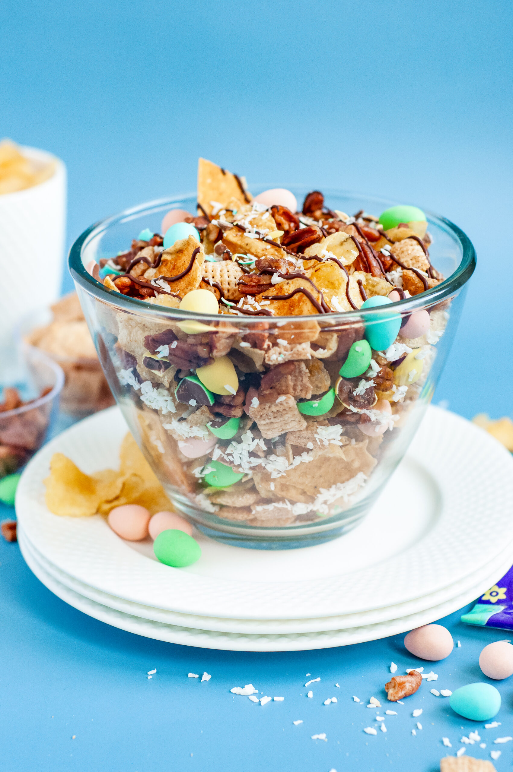 Bowl of garden party snack mix with candied pecans, kettle chips, pastel candies, and a dark chocolate drizzle, sprinkled with flaky sea salt.
