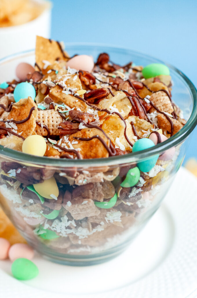 Bowl of garden party snack mix with candied pecans, kettle chips, pastel candies, and a dark chocolate drizzle, sprinkled with flaky sea salt.