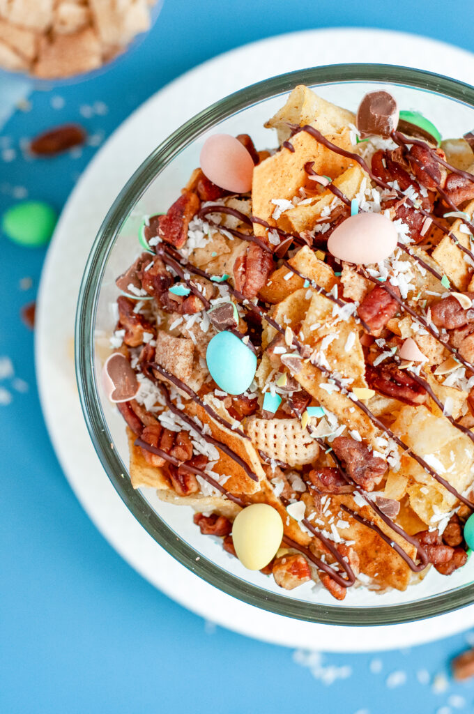Bowl of garden party snack mix with candied pecans, kettle chips, pastel candies, and a dark chocolate drizzle, sprinkled with flaky sea salt.