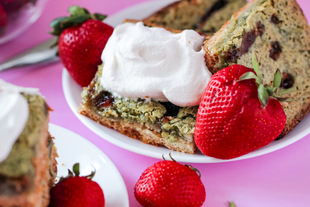 A slice of gooey butter cake with a green matcha filling swirled with strawberry jam, topped with fresh strawberries and whipped cream on a white plate.