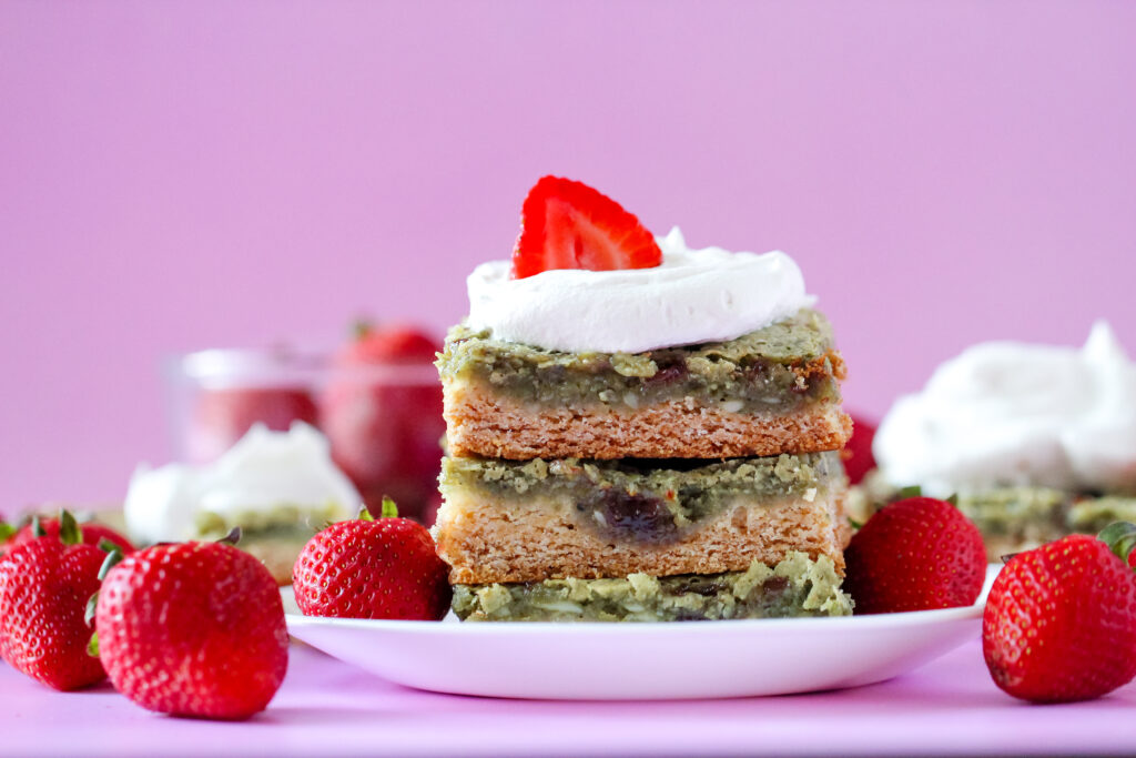 A slice of gooey butter cake with a green matcha filling swirled with strawberry jam, topped with fresh strawberries and whipped cream on a white plate.
