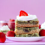 A slice of gooey butter cake with a green matcha filling swirled with strawberry jam, topped with fresh strawberries and whipped cream on a white plate.