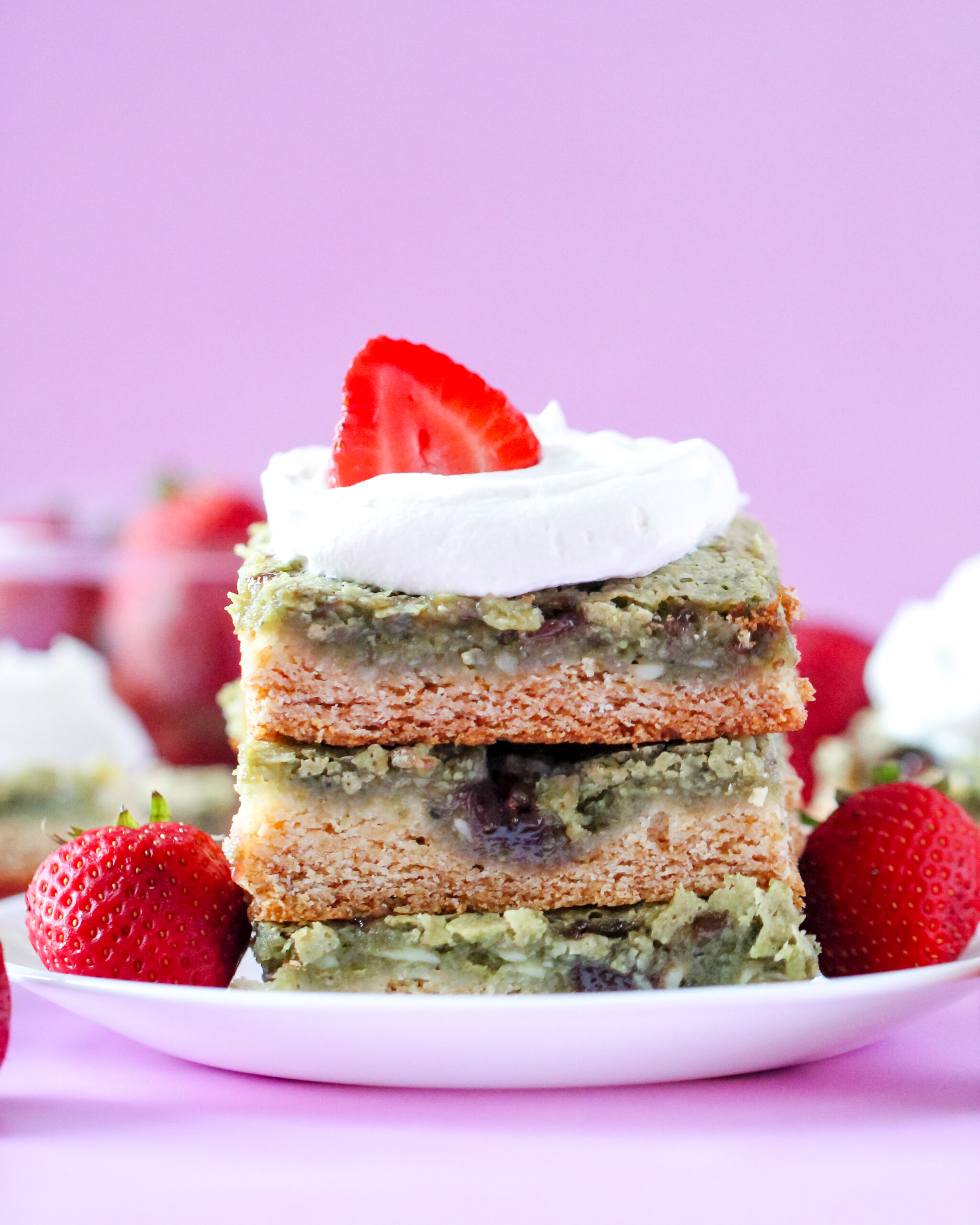 A slice of gooey butter cake with a green matcha filling swirled with strawberry jam, topped with fresh strawberries and whipped cream on a white plate.