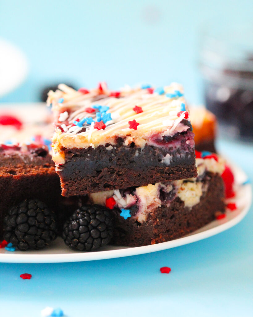 Patriotic cheesecake brownies with red and blue jam swirls, white chocolate drizzle, and star-shaped sprinkles on top.