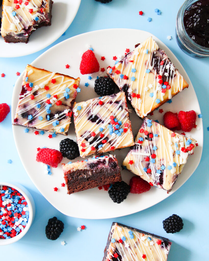 Patriotic cheesecake brownies with red and blue jam swirls, white chocolate drizzle, and star-shaped sprinkles on top.