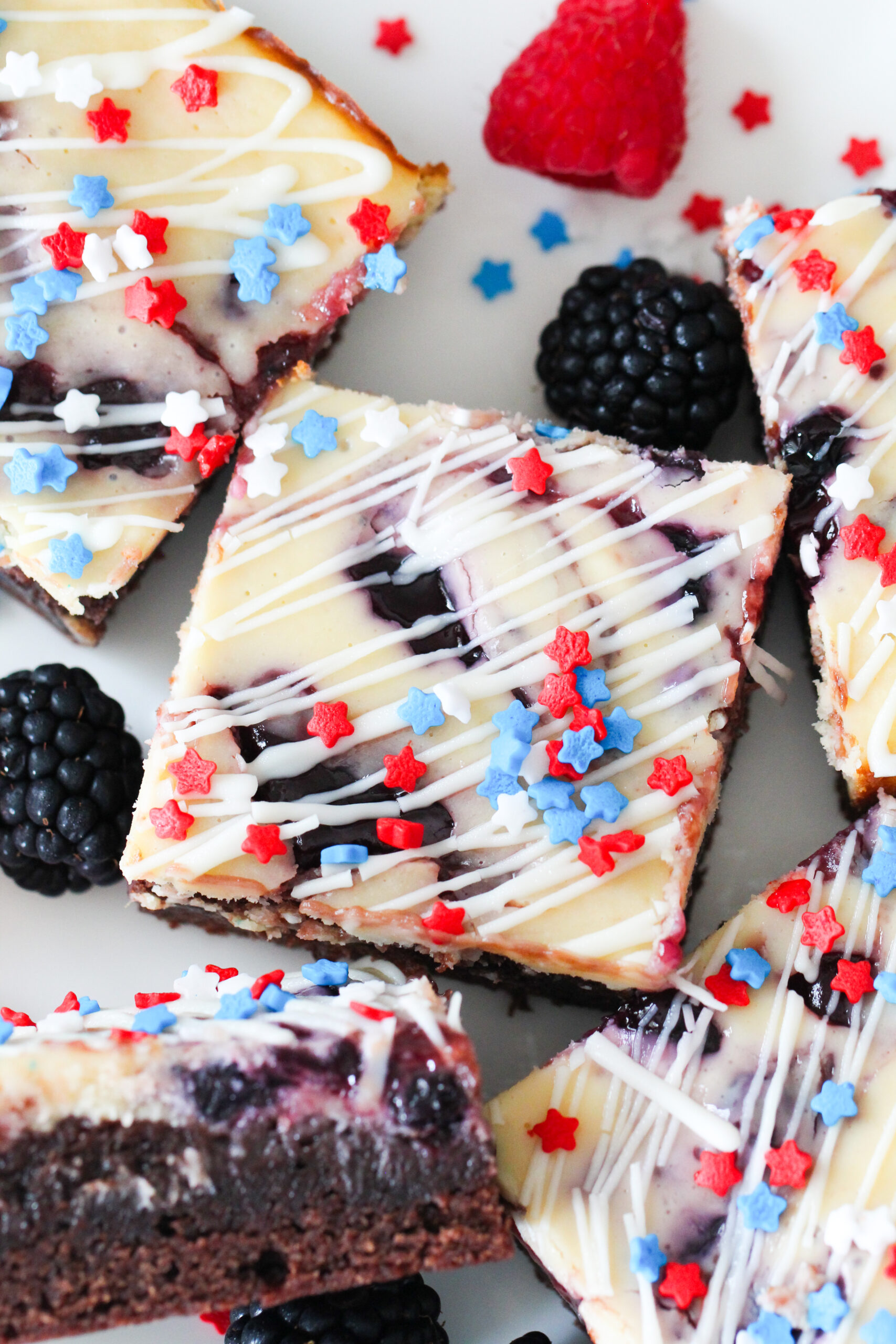 Patriotic cheesecake brownies with red and blue jam swirls, white chocolate drizzle, and star-shaped sprinkles on top.