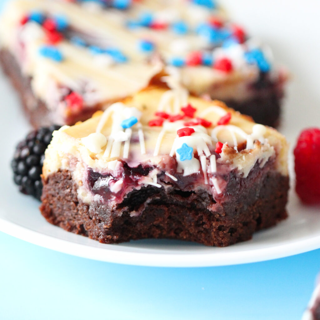 Patriotic cheesecake brownies with red and blue jam swirls, white chocolate drizzle, and star-shaped sprinkles on top.