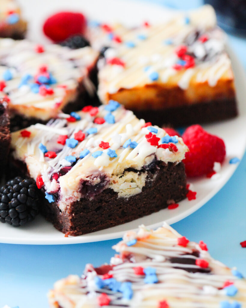 Patriotic cheesecake brownies with red and blue jam swirls, white chocolate drizzle, and star-shaped sprinkles on top.