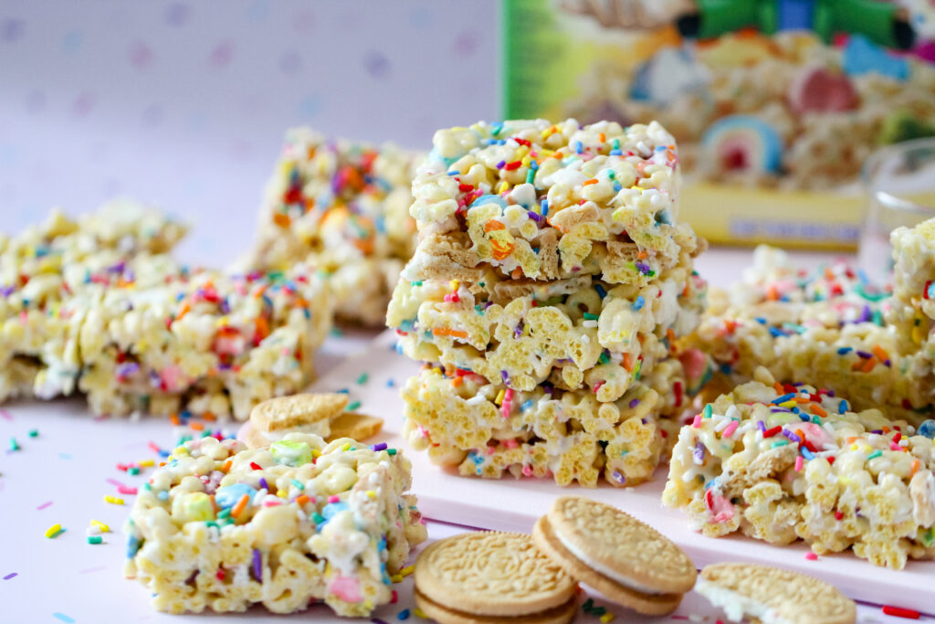 Colorful Lucky Charms cereal bars topped with rainbow sprinkles, Golden Oreo pieces, and melted white chocolate in a 13x9 pan.
