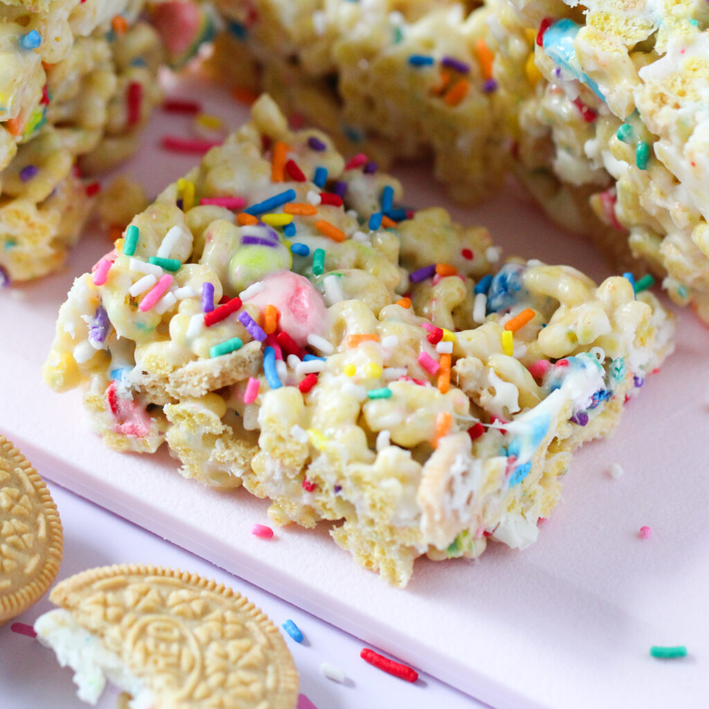Colorful Lucky Charms cereal bars topped with rainbow sprinkles, Golden Oreo pieces, and melted white chocolate in a 13x9 pan.