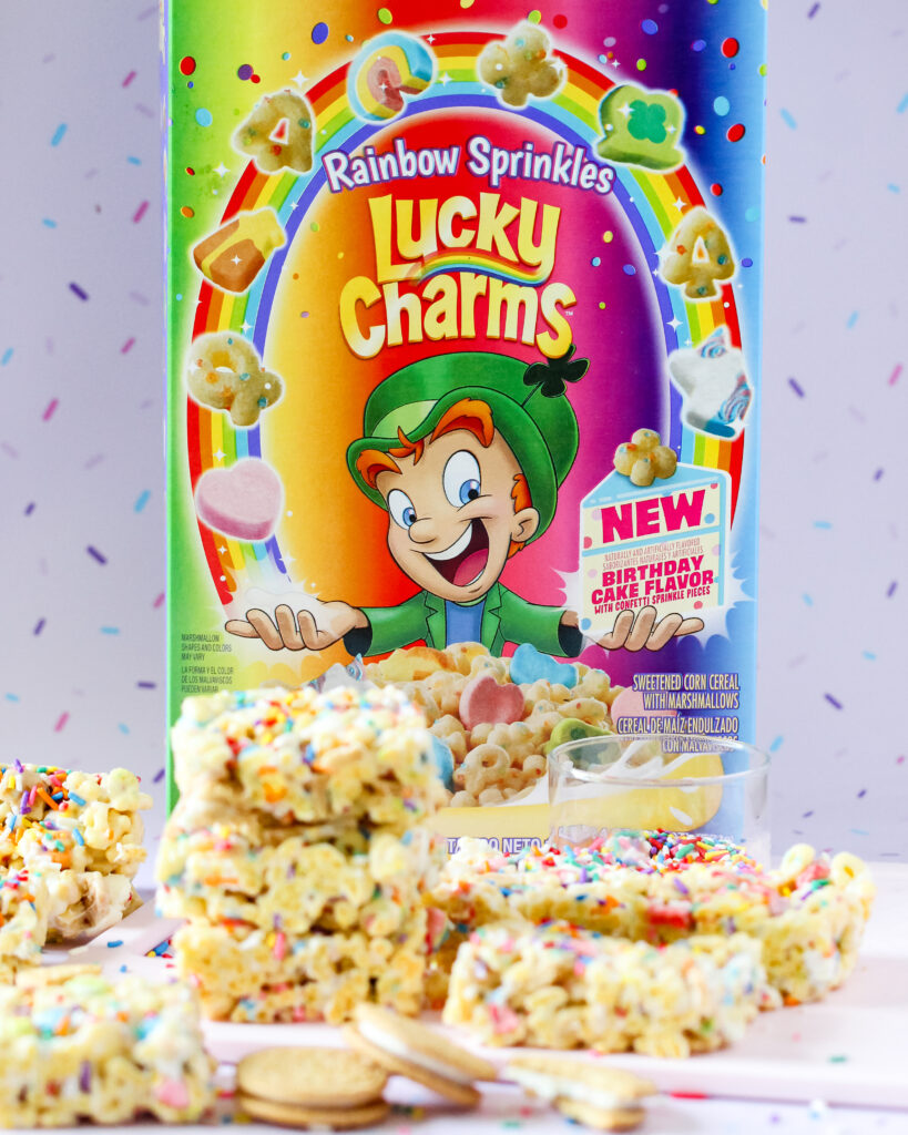 Colorful Lucky Charms cereal bars topped with rainbow sprinkles, Golden Oreo pieces, and melted white chocolate in a 13x9 pan.