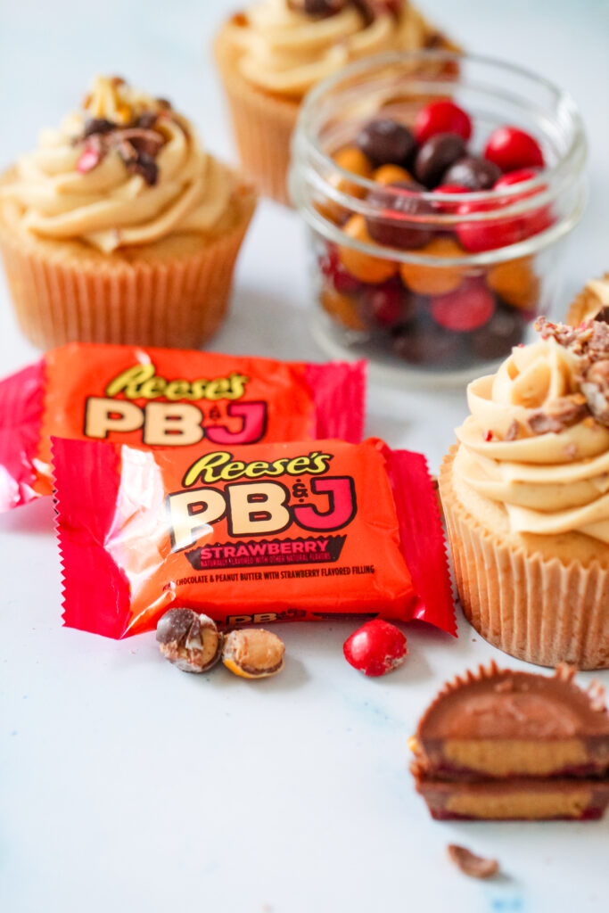 Strawberry PB&J cupcakes topped with fluffy peanut butter frosting, chopped Reese’s PB&J Cups, and colorful candy pieces.