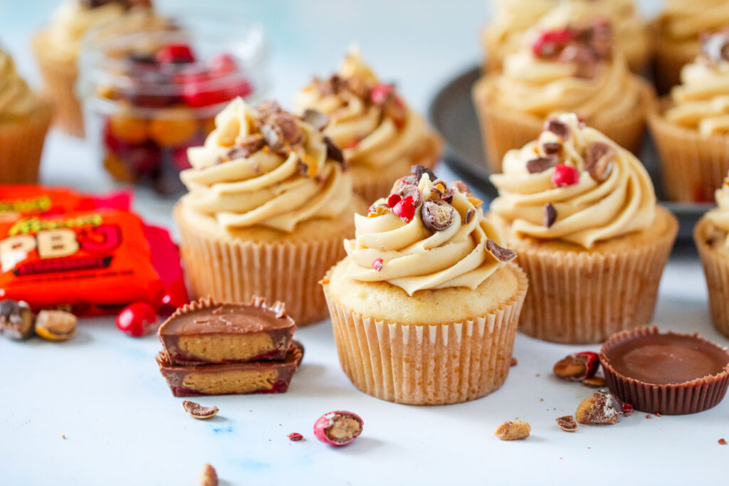Strawberry PB&J cupcakes topped with fluffy peanut butter frosting, chopped Reese’s PB&J Cups, and colorful candy pieces.