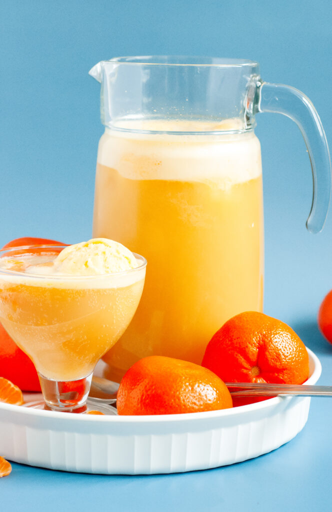 A punch bowl filled with orange sherbet punch, garnished with orange slices and cherries, surrounded by bright summer party decor.
