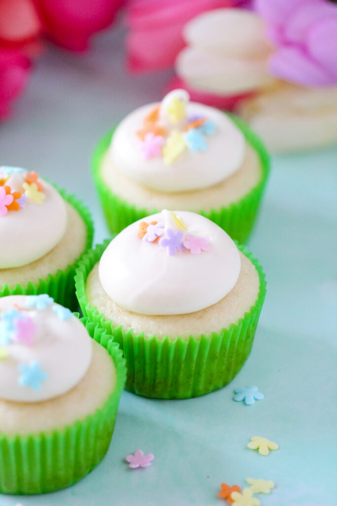 Almond cupcakes topped with honey cream cheese frosting, spring sprinkles, and a drizzle of golden honey on a white plate.