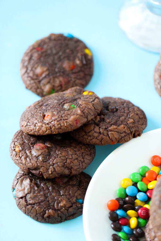 A stack of rich, fudgy brownie cookies studded with mini M&Ms, sitting on a parchment-lined wire rack with colorful candies scattered around.