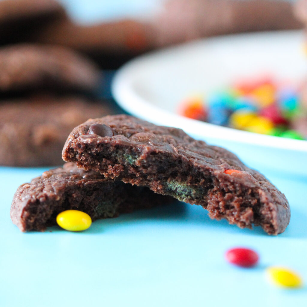 A stack of rich, fudgy brownie cookies studded with mini M&Ms, sitting on a parchment-lined wire rack with colorful candies scattered around.