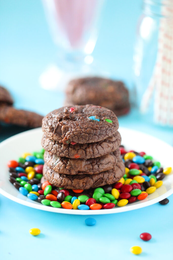 A stack of rich, fudgy brownie cookies studded with mini M&Ms, sitting on a parchment-lined wire rack with colorful candies scattered around.